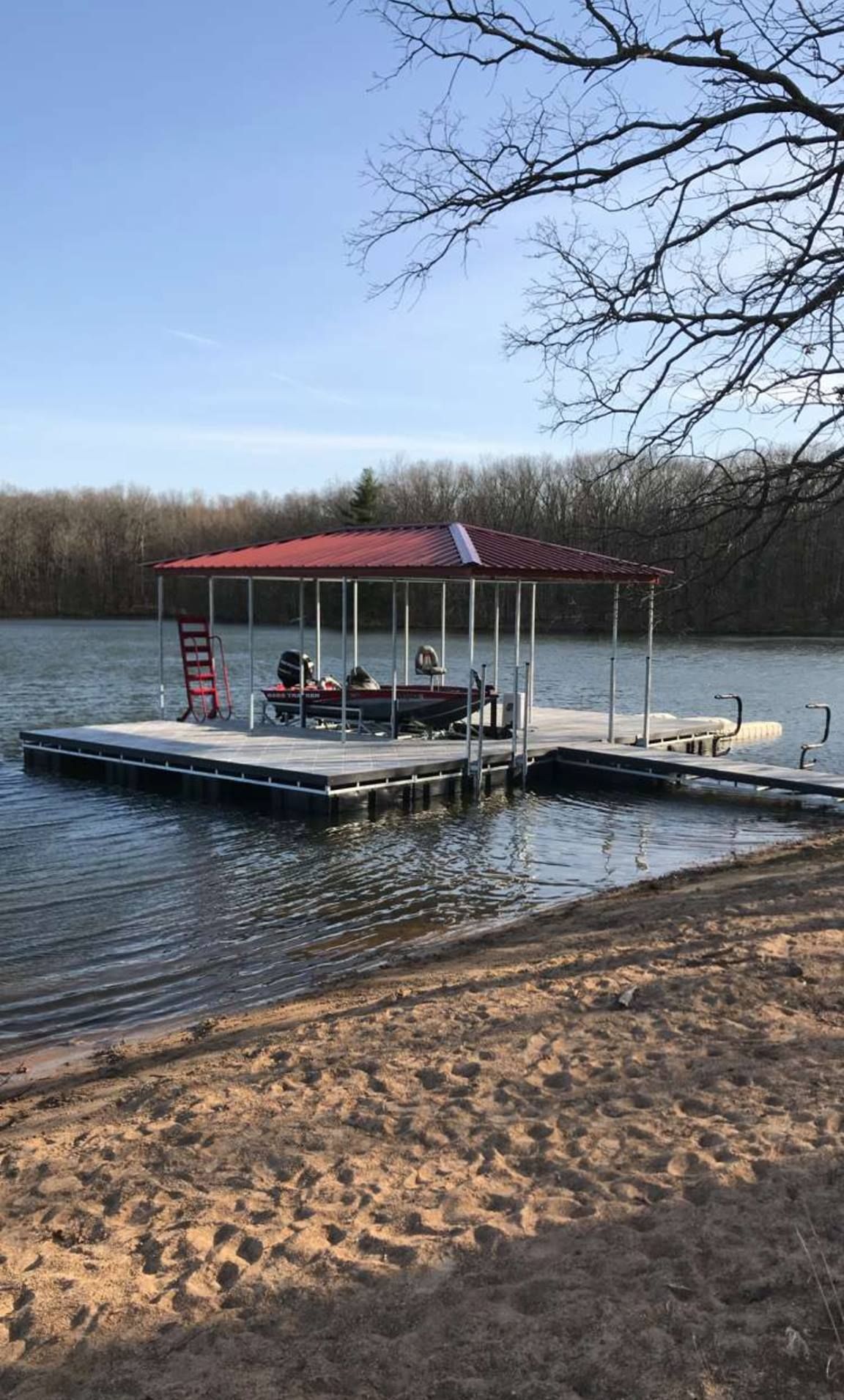 Covered dock with a red roof on a calm lake, with a small boat and sandy shore in the foreground