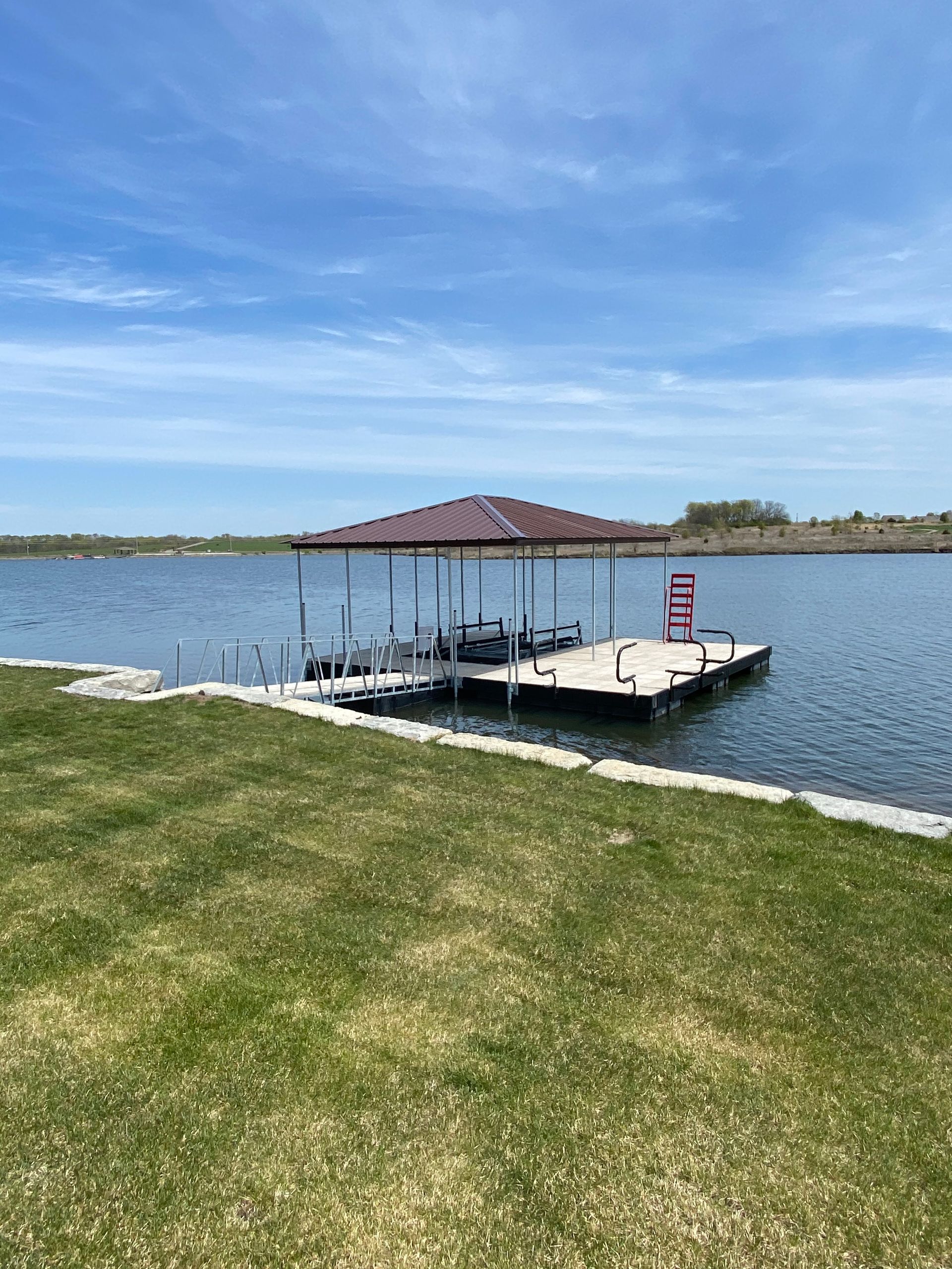 Covered dock on a calm lake beside a grassy shore under a blue sky