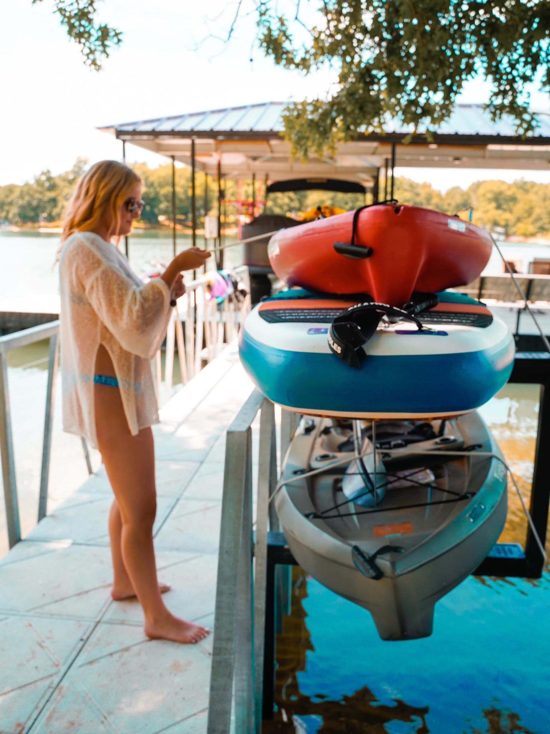 Woman standing beside stacked kayaks on a dock next to clear blue water.