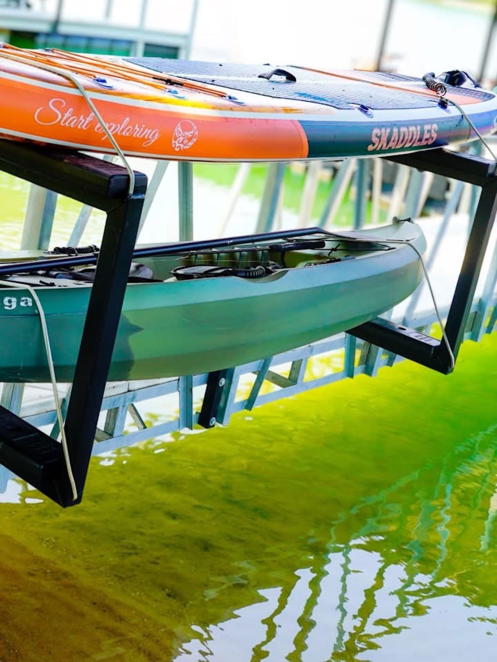 Two kayaks stacked on a rack above green water, with reflections in the canal below.