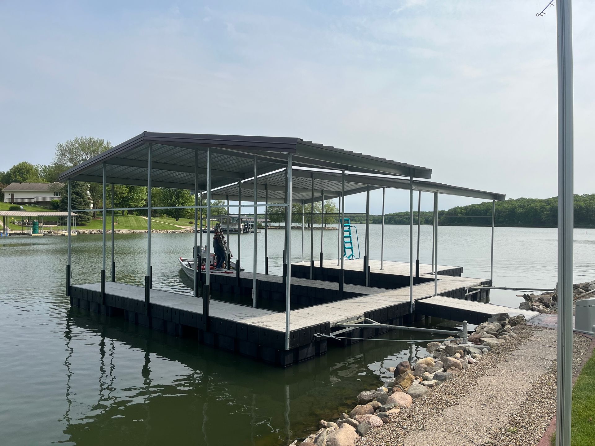 Covered dock on a calm lake with a metal roof and rocky shoreline under a cloudy sky