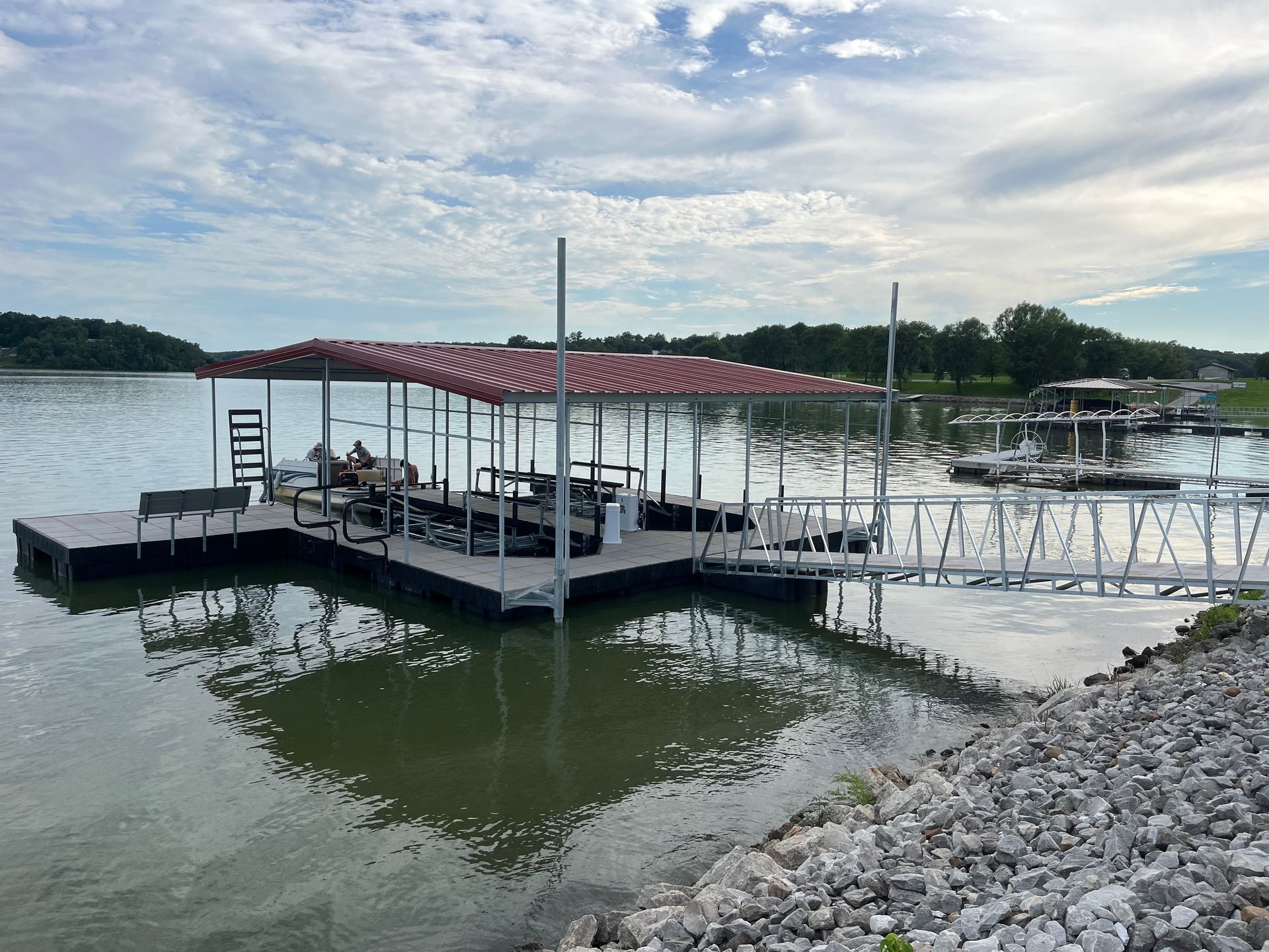 Covered dock on a calm lake with a red roof, floating walkway, and rocky shoreline under a cloudy sky