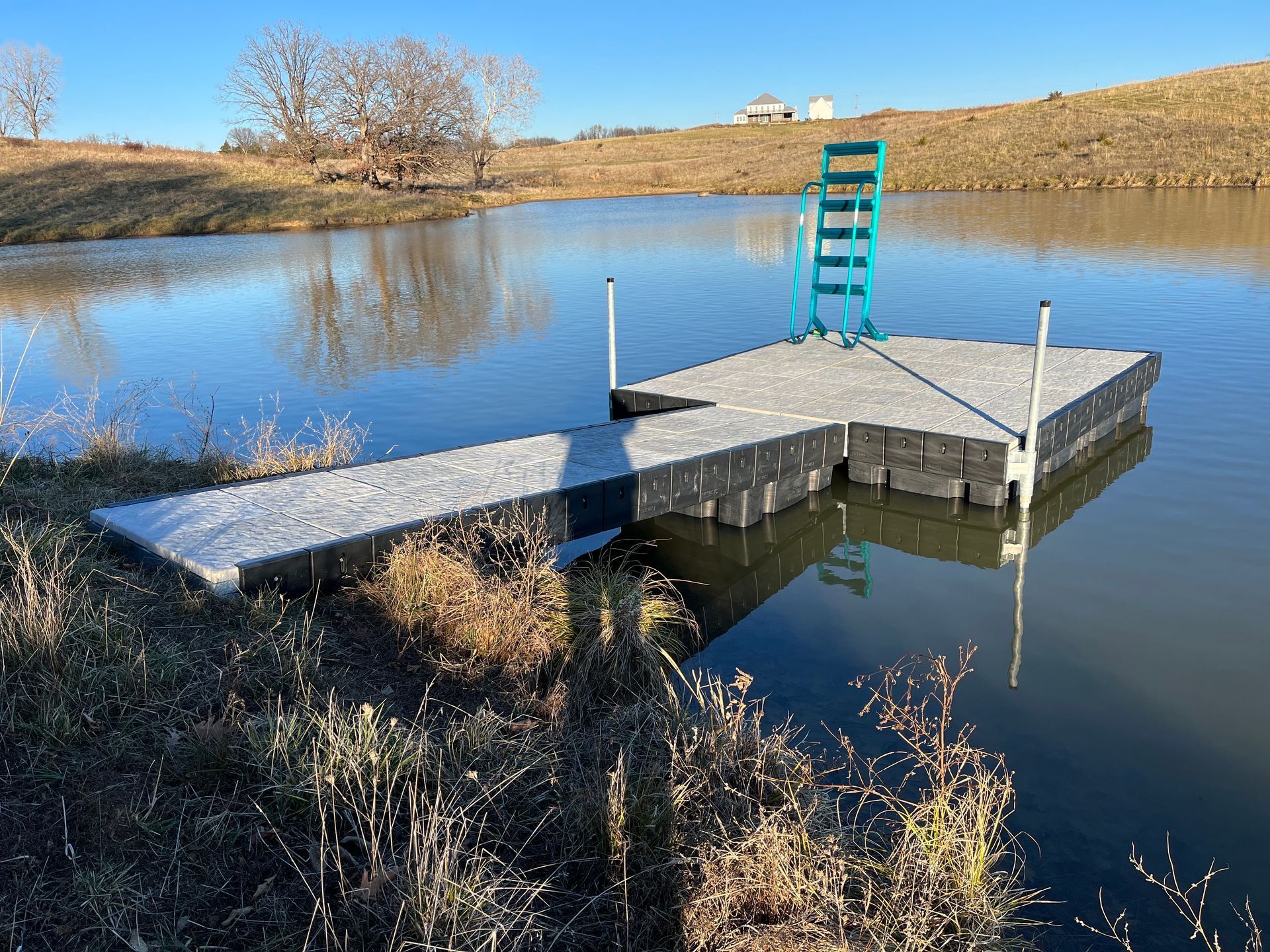 Small dock with a green ladder on a calm pond, surrounded by dry grass and bare trees under a clear sky.