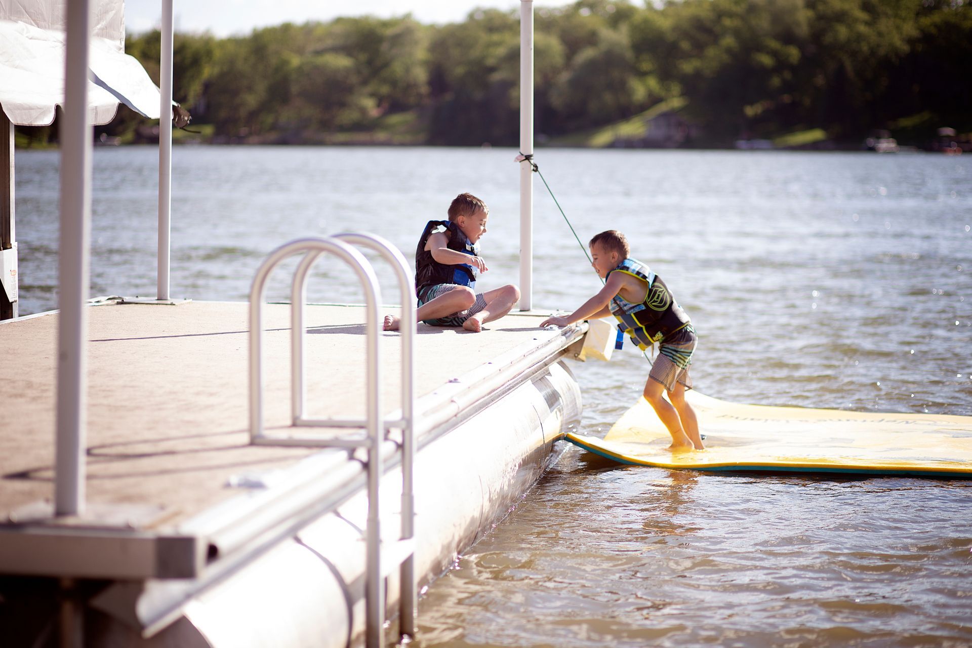 Person climbing onto a dock from a yellow board while another sits on the dock by the lake