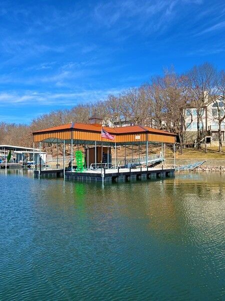 Covered dock with boats on a calm lake beside a wooded shoreline under a blue sky