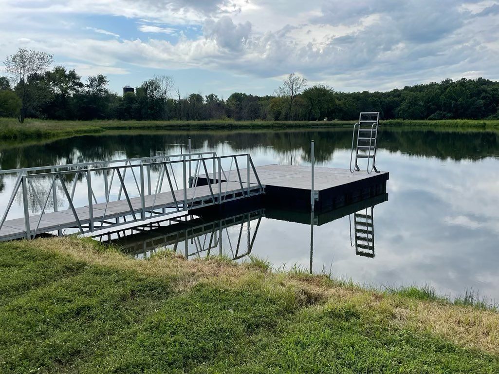 Floating dock with chairs on a calm pond, surrounded by green grass and trees under a cloudy sky