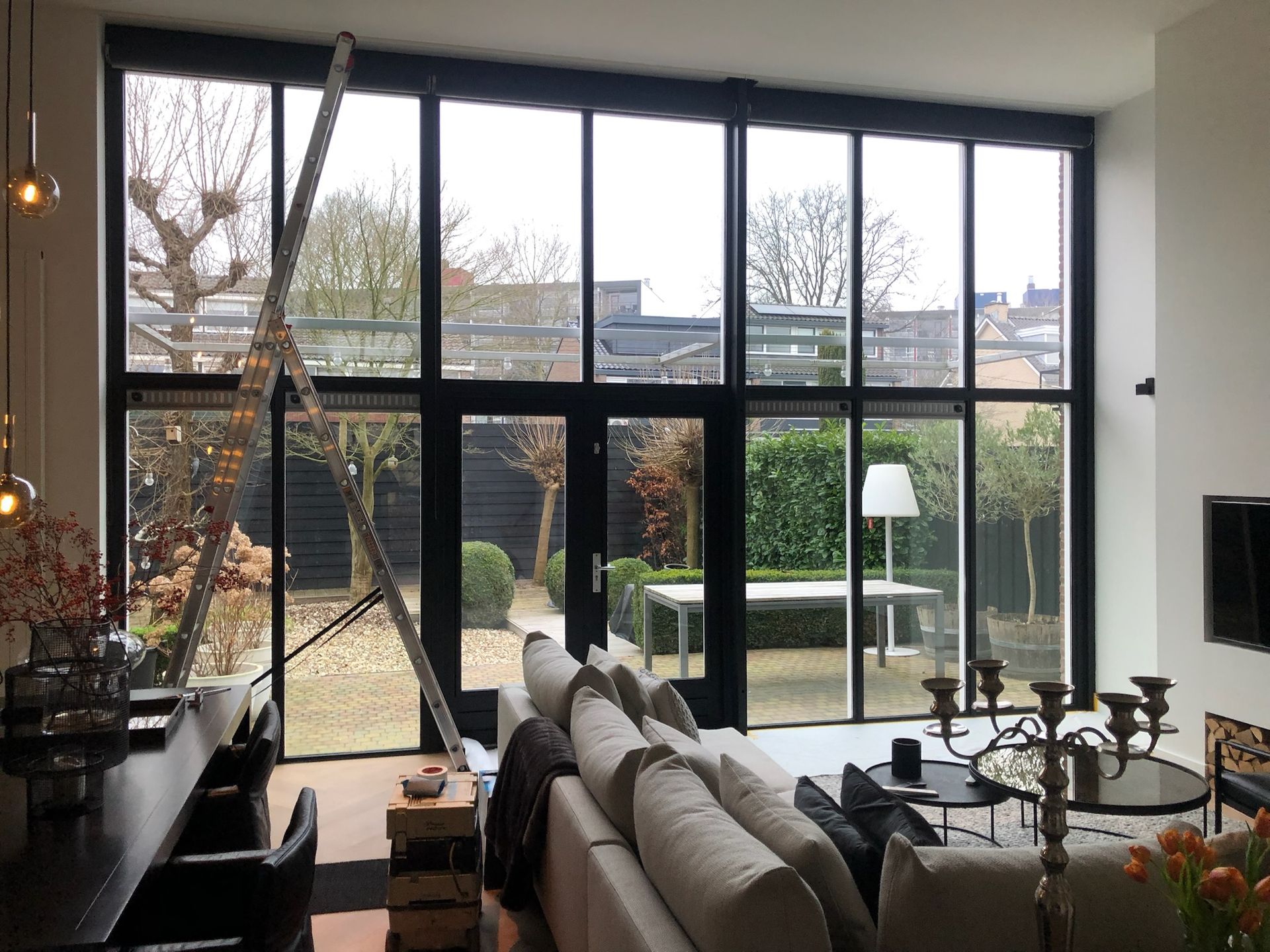 Interieur van een moderne woonkamer met grote zwarte ramen die uitkijken op een achtertuin met een tafel en planten.