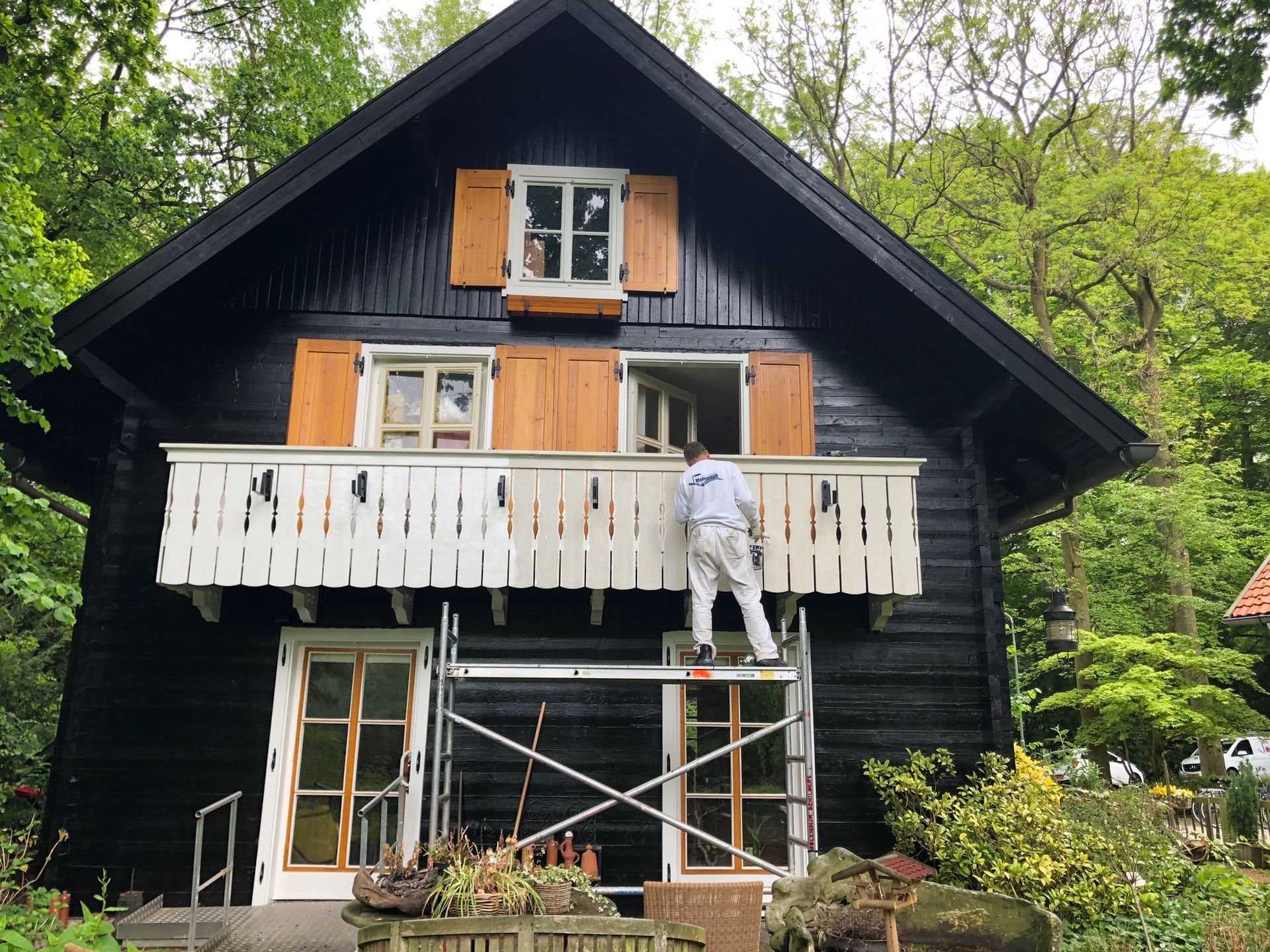 Persoon die een witte leuning schildert op een zwarte hut met oranje luiken, staand op een steiger.