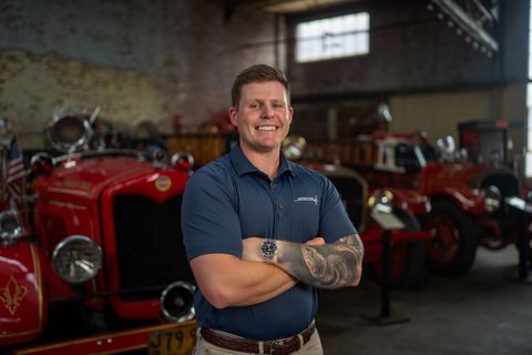 Smiling man with tattooed arms standing in front of vintage red cars in a garage