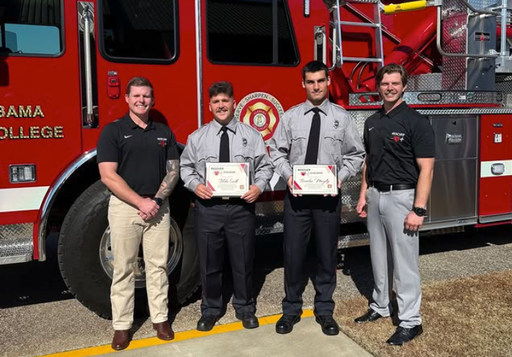 Two men holding certificates beside a red fire truck with two firefighters