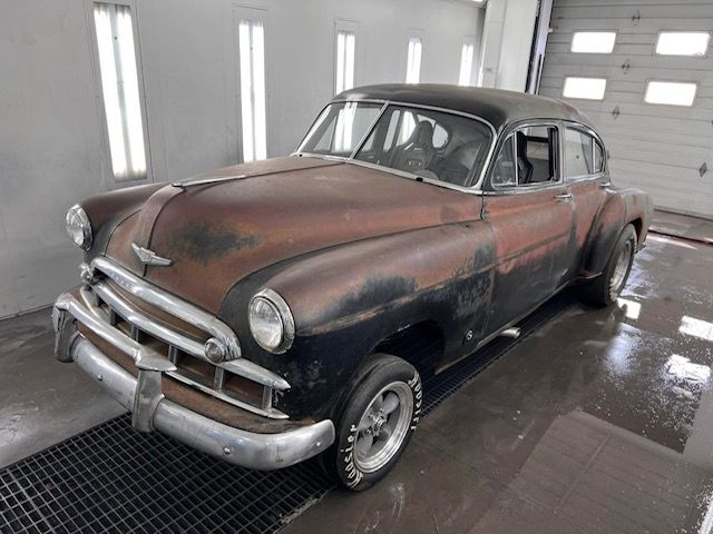 A rusty classic Chevrolet car in a workshop, black and brown paint, chrome bumper, wide wheels.