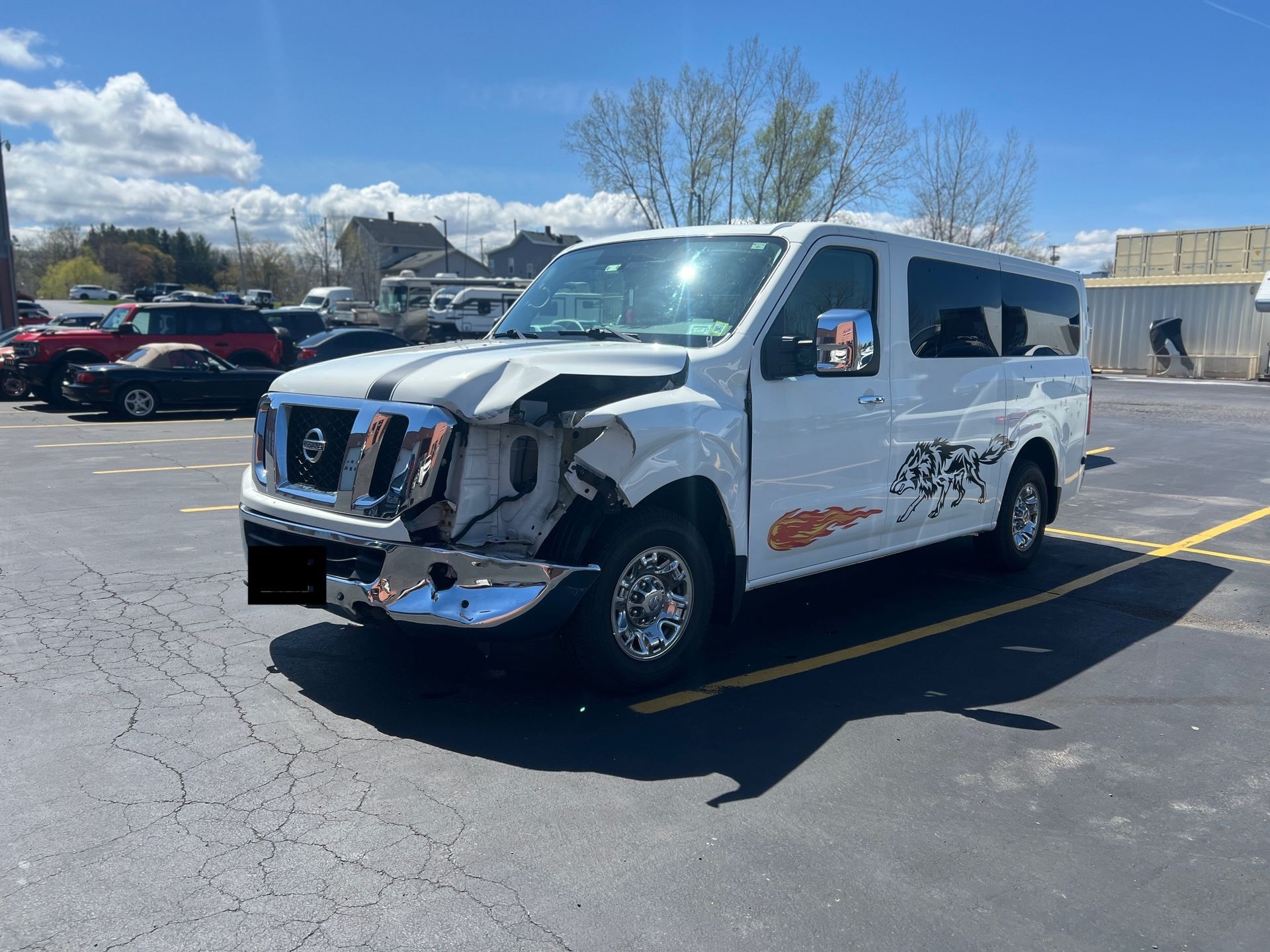 White shuttle van with front-end damage parked in a sunny lot, with a blue sky and buildings in the background