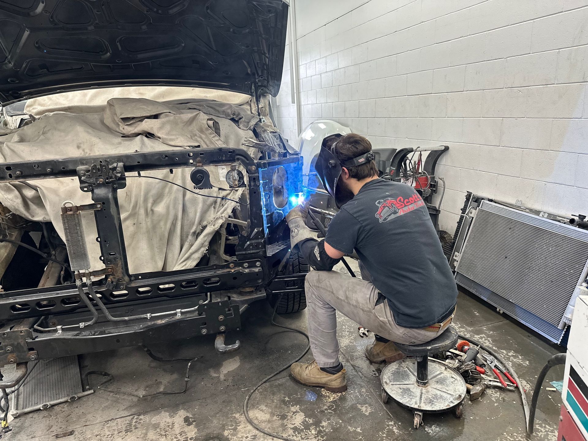 Mechanic welding the front of a damaged car in a garage, bright blue sparks visible.