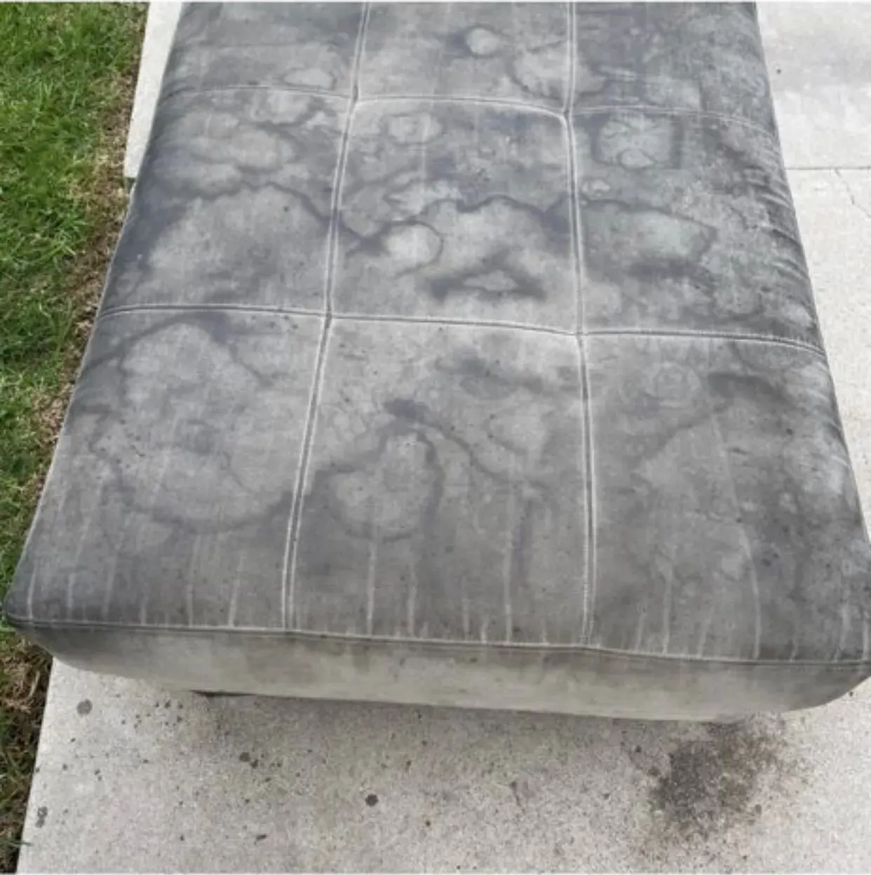 Worn gray outdoor cushion or mattress on concrete beside grass