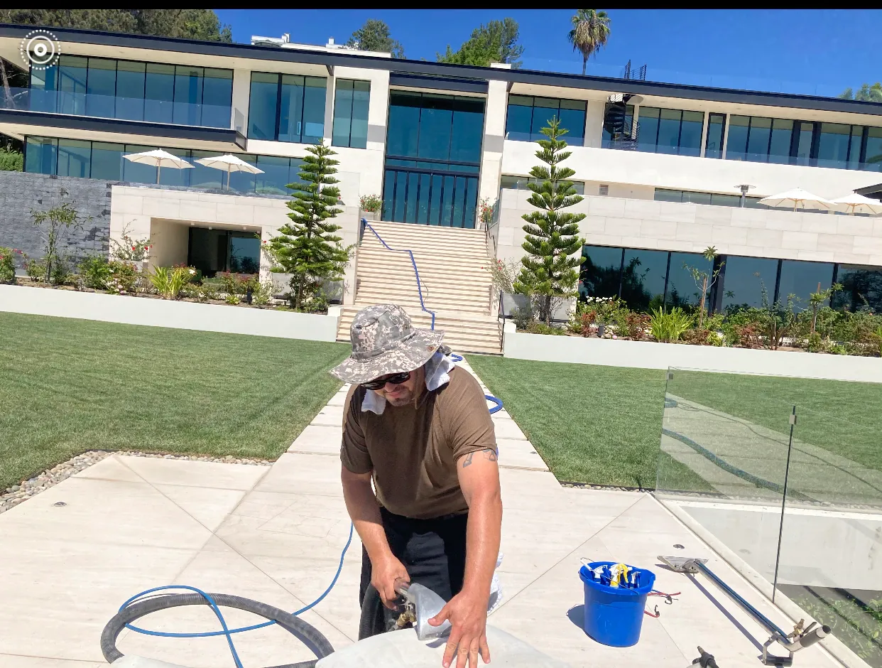 Person in a sun hat cleaning a patio with a hose in front of a modern house.