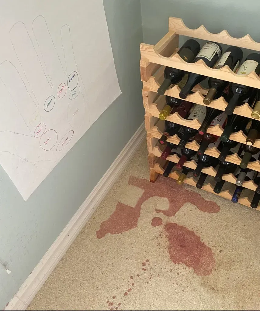 Spilled red liquid on beige carpet beside a wooden wine rack and a white wall with paper notes