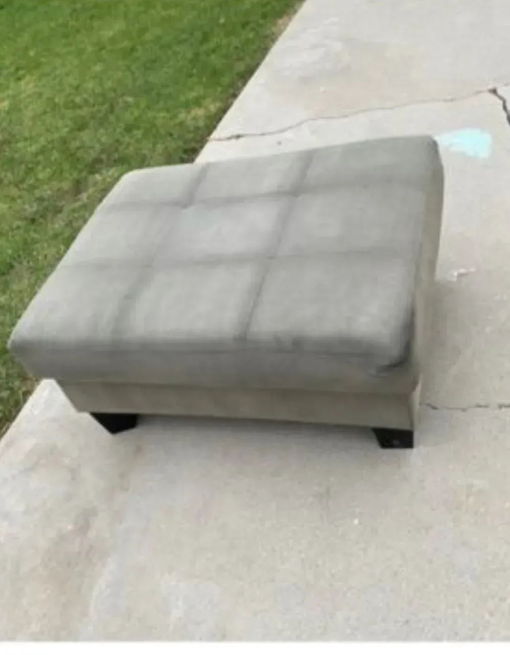 Gray upholstered ottoman on a concrete sidewalk beside a grassy lawn