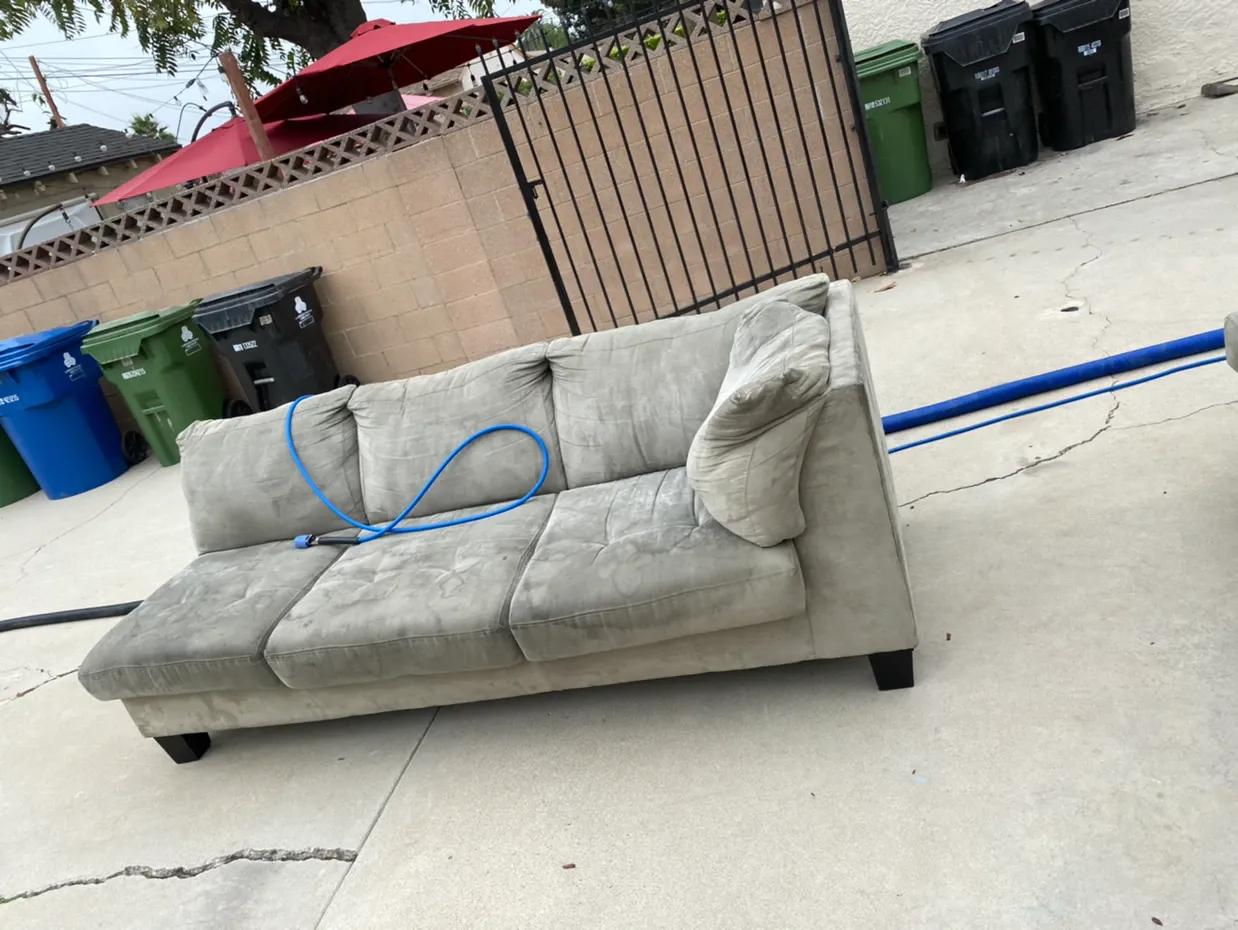 Gray couch on a concrete driveway near trash bins and a tan fence, with a blue hose draped across it.