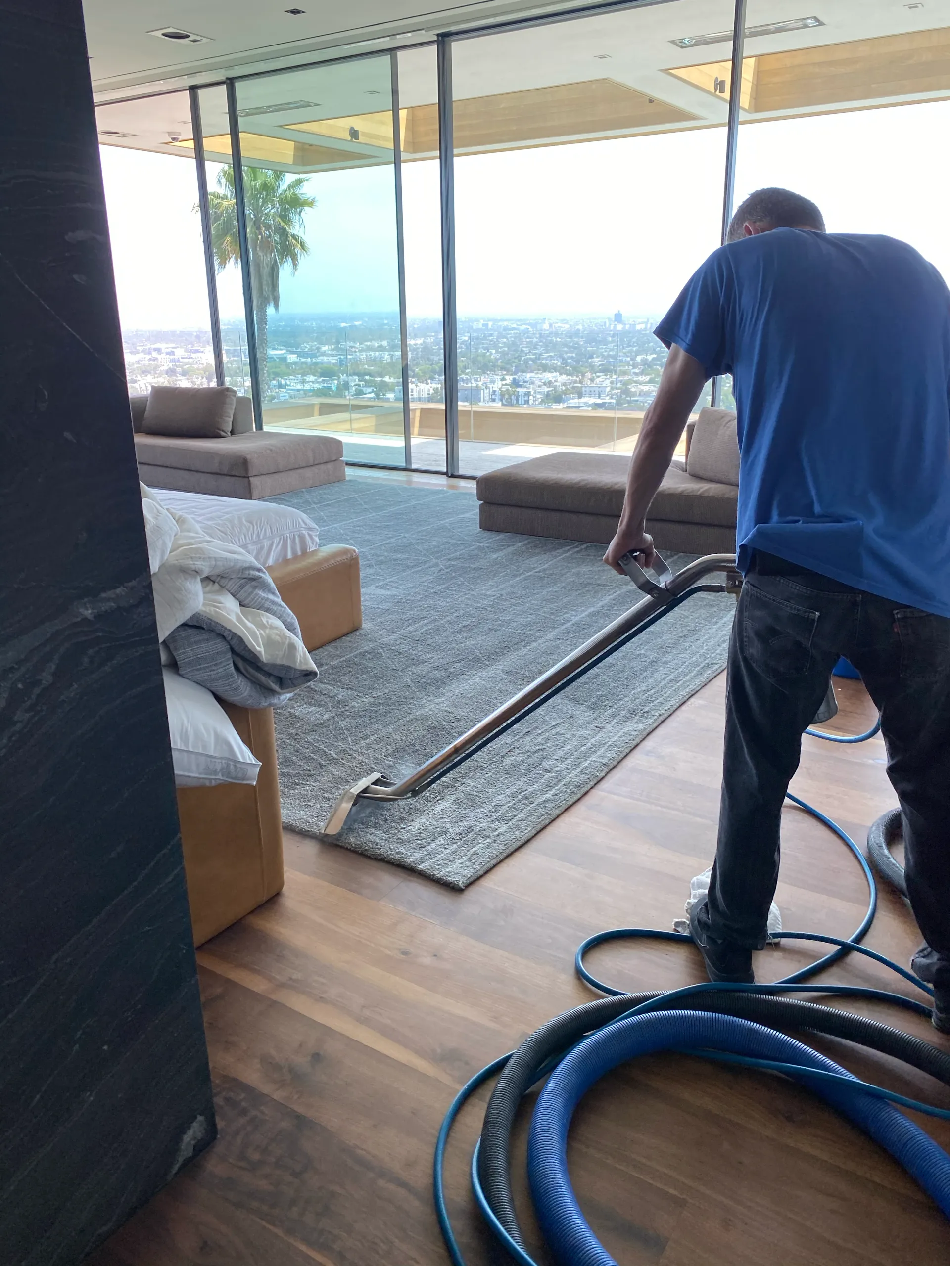 Worker vacuuming dust in a modern living room with large windows and city view.
