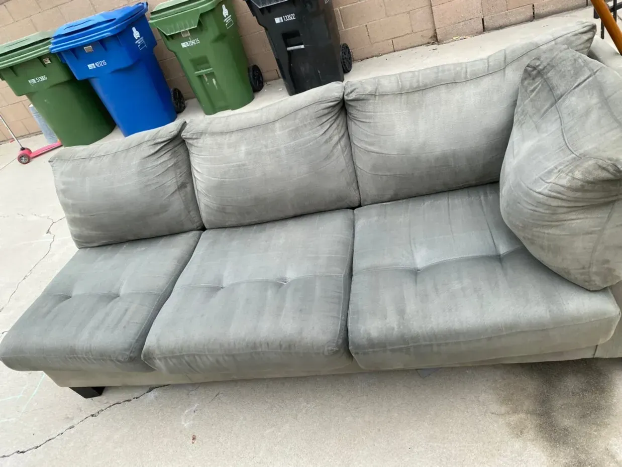Gray three-seat sofa on a sidewalk beside blue and green trash bins.