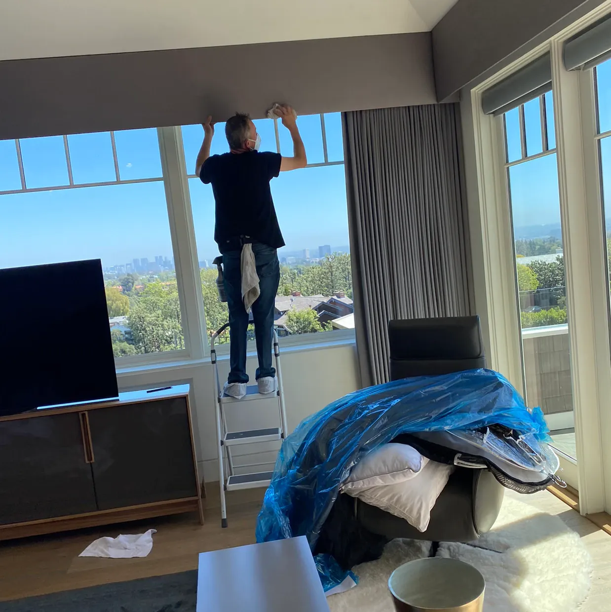 Person standing on a ladder by a large window, adjusting the top blinds in a bright room.