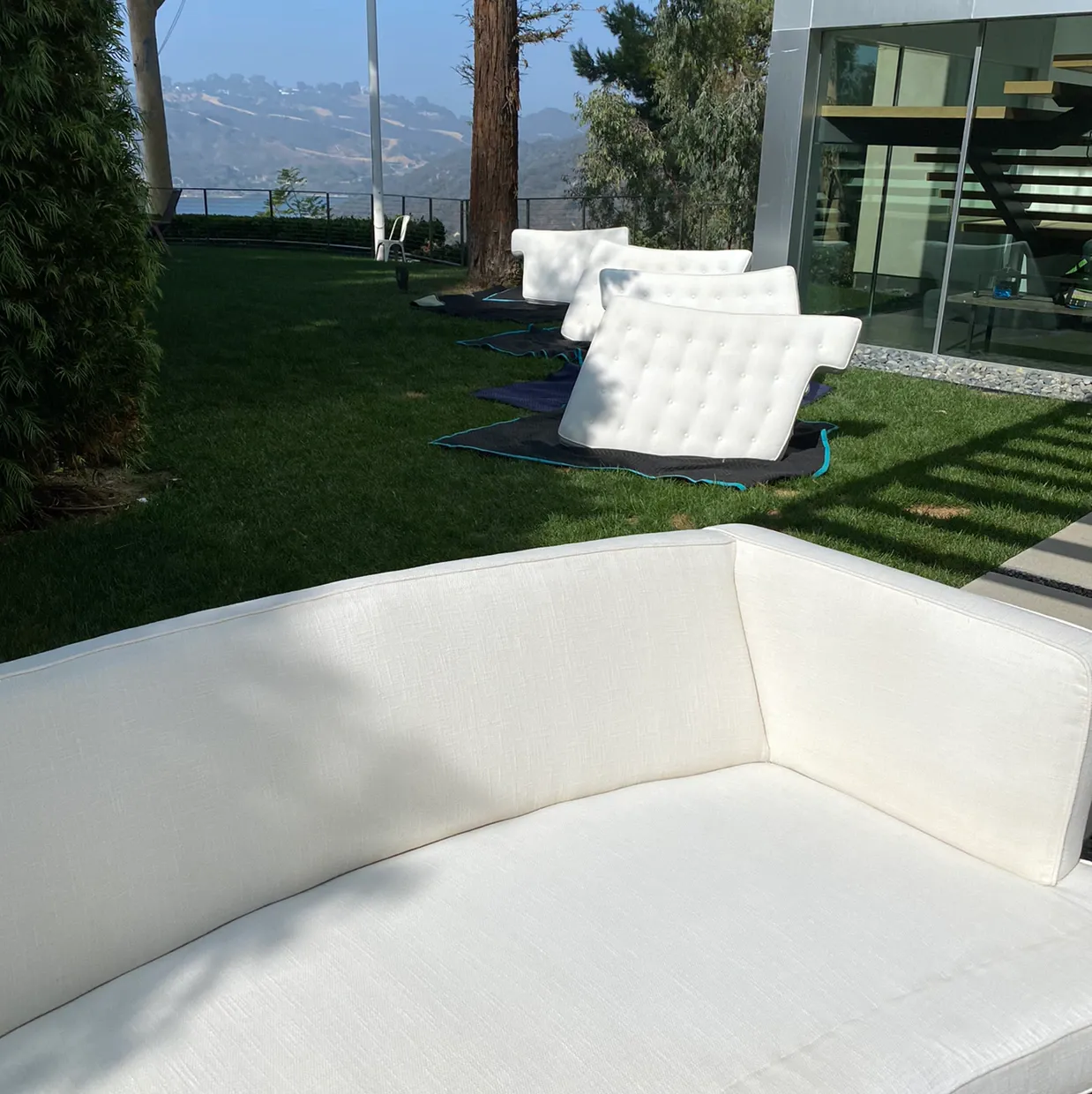 White patio sofa on a lawn beside a modern glass house, with more white chairs and mountain views in the background