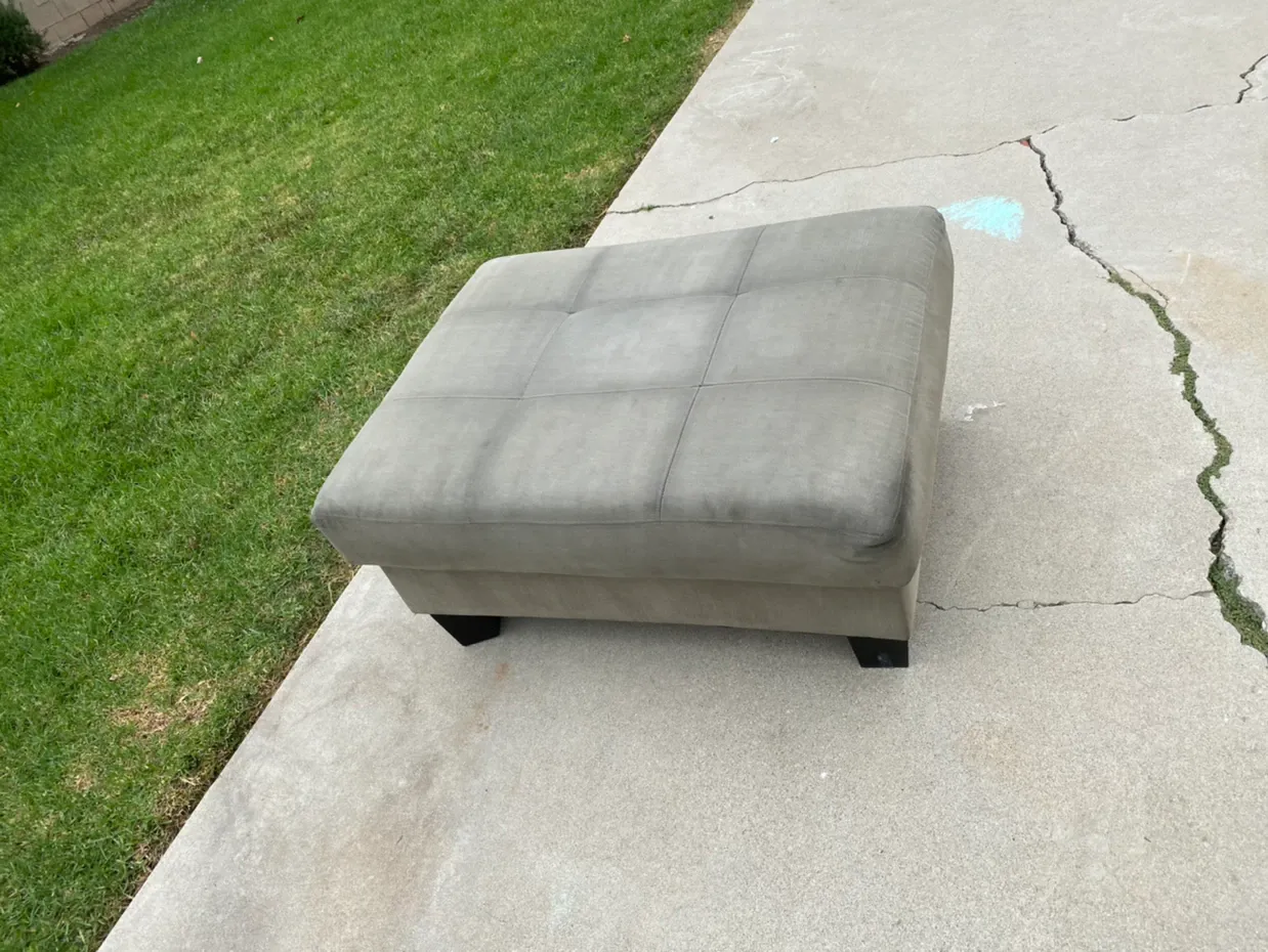 Gray ottoman on a concrete driveway beside grass, near a crack in the pavement