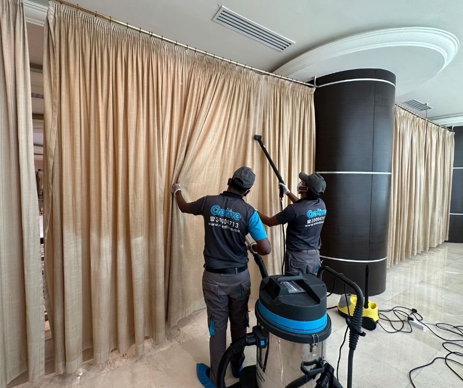 Two workers installing beige curtains in a bright indoor room, with a vacuum and tools nearby