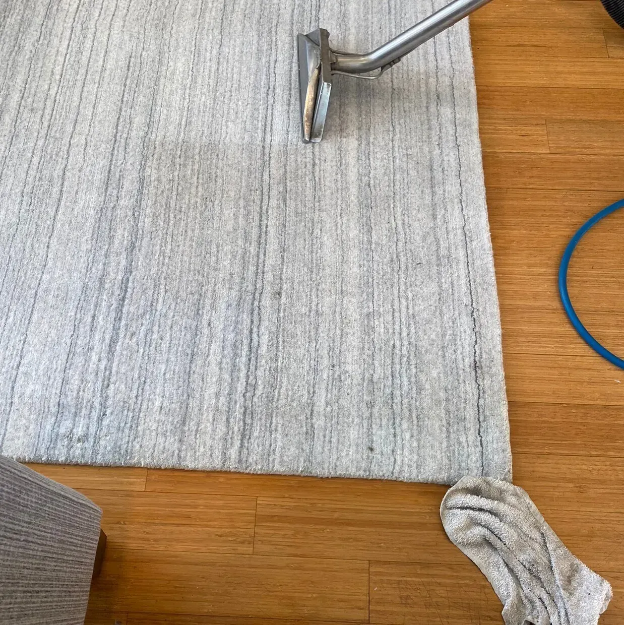 Gray rug being vacuumed on a wooden floor, with a blue cord and a crumpled cloth nearby.