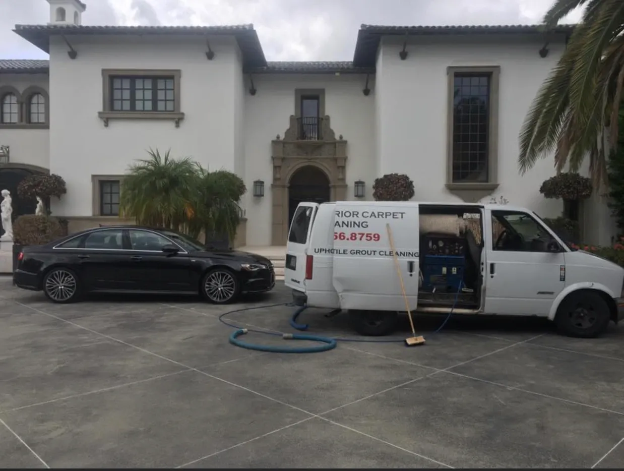 White service van and black sedan parked in front of a white stucco house with an open garage and hose on driveway