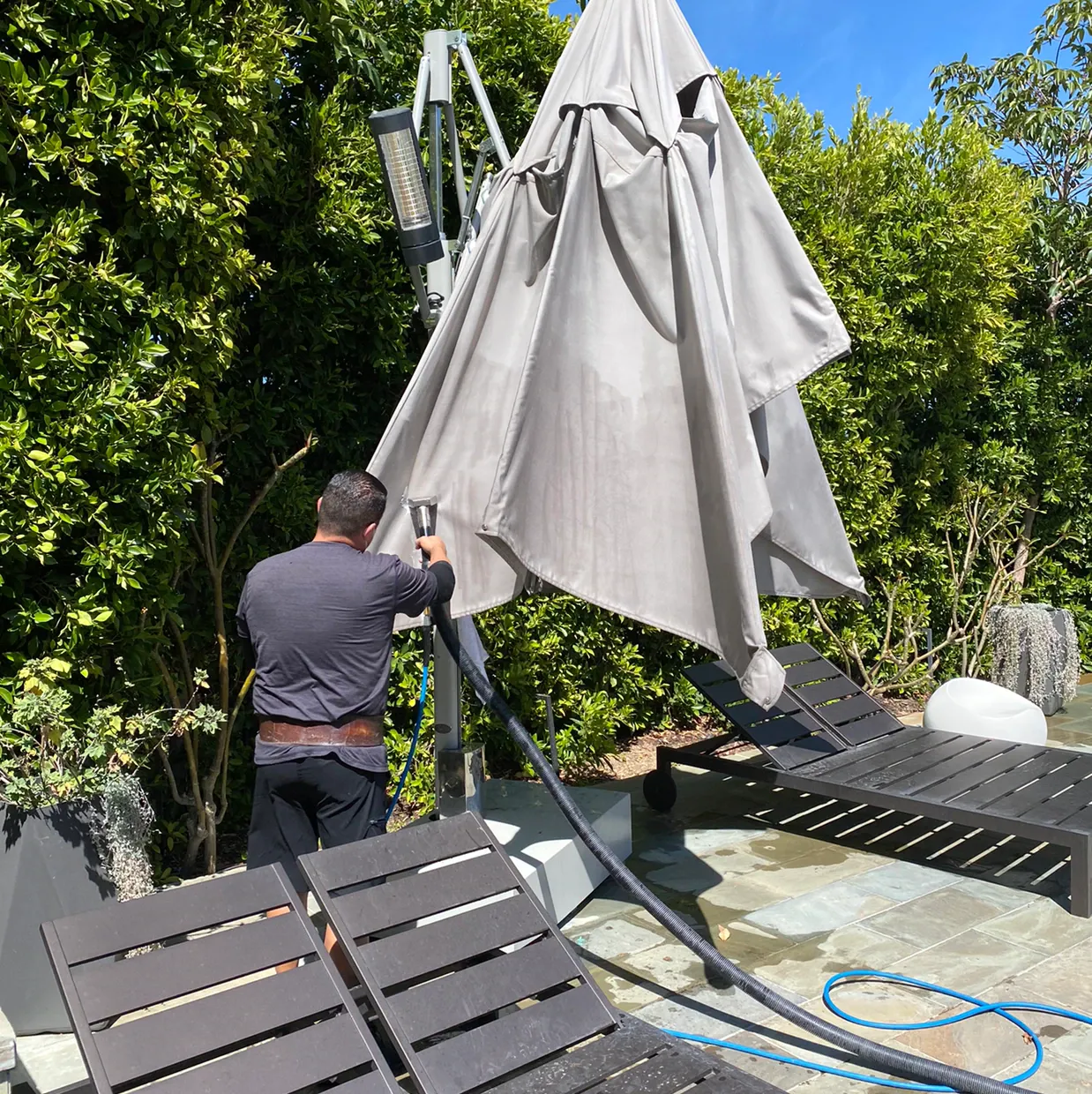 Person lowering a large gray patio umbrella beside a pool, with lounge chairs and greenery nearby