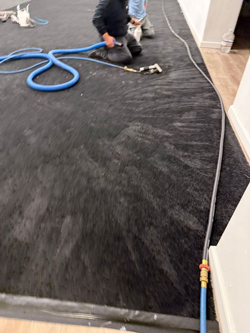Worker vacuuming black carpet during installation, with blue hose in a room under construction