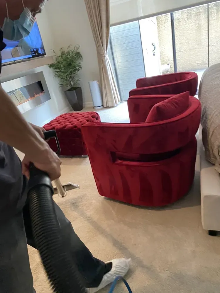 Living room with red velvet chairs, a beige sofa, and a person holding a vacuum cleaner.