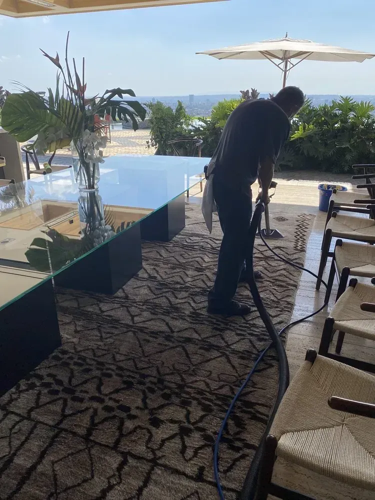 Person vacuuming carpet on a shaded patio beside a pool with ocean view