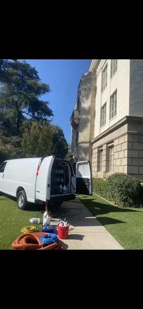 White van parked by a stone building; an open rear door reveals a person inside, with boxes on the grass.