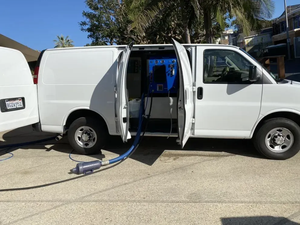 White utility van with side door open, showing equipment inside and a blue hose on a driveway.