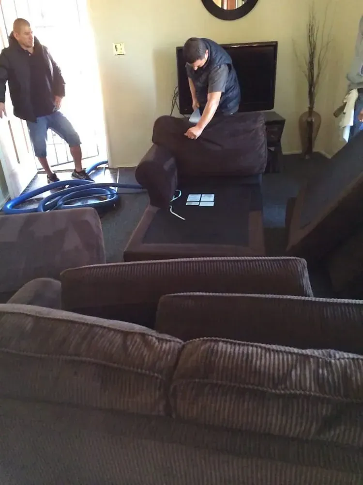 Living room with two people moving a dark brown chair, sofas and a TV in the background