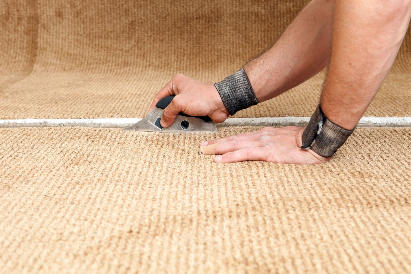 Person cutting carpet with a utility knife, measuring along a white line on a tan floor.