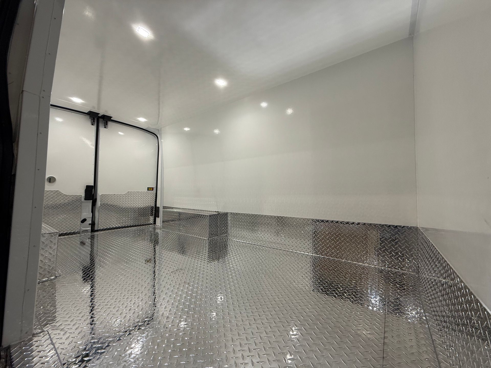 Interior of a cargo trailer with white walls, ceiling lights, and diamond plate metal flooring.