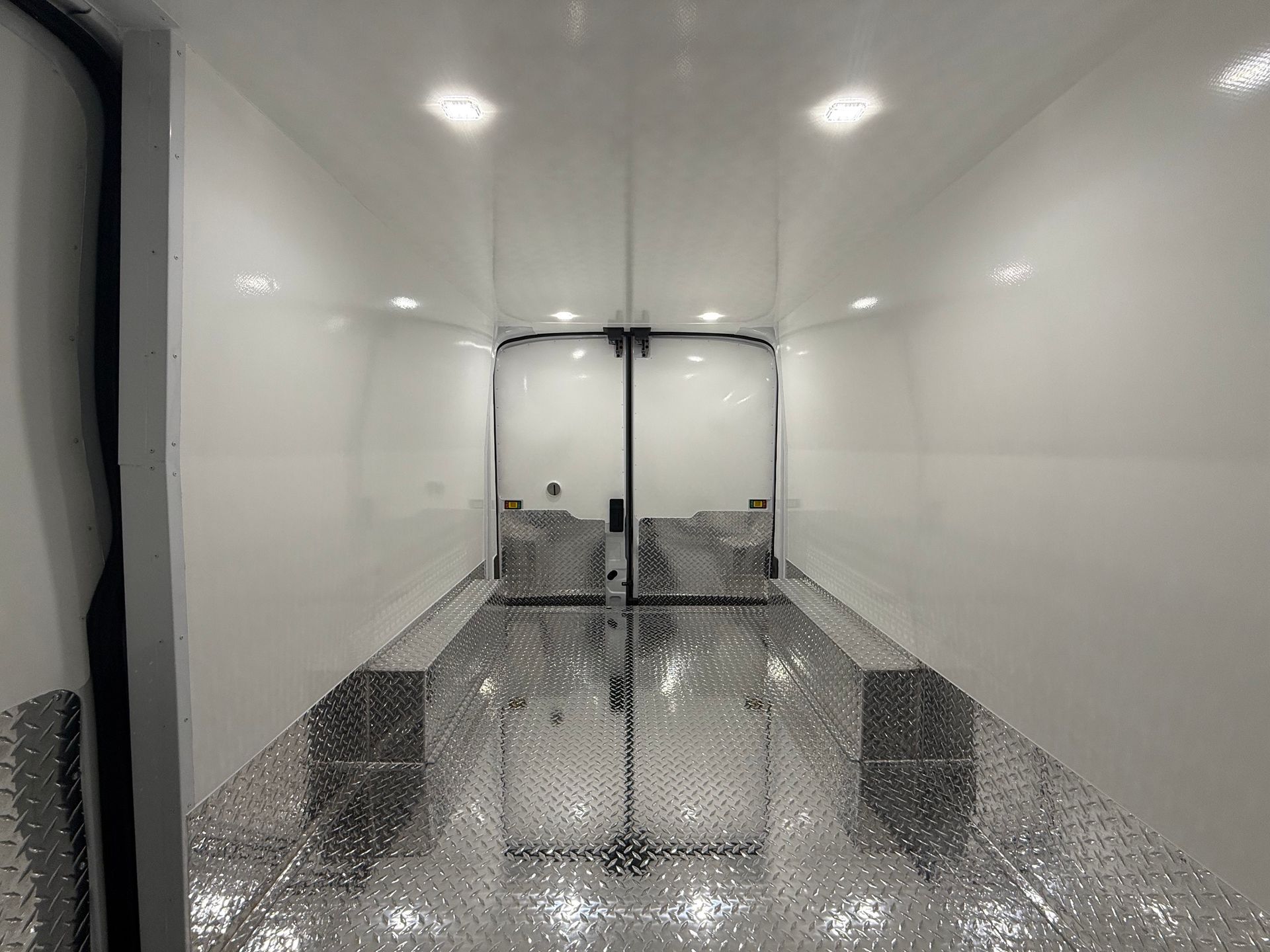 An empty cargo trailer interior with white walls, ceiling LED lights, and a diamond-plate metal floor.