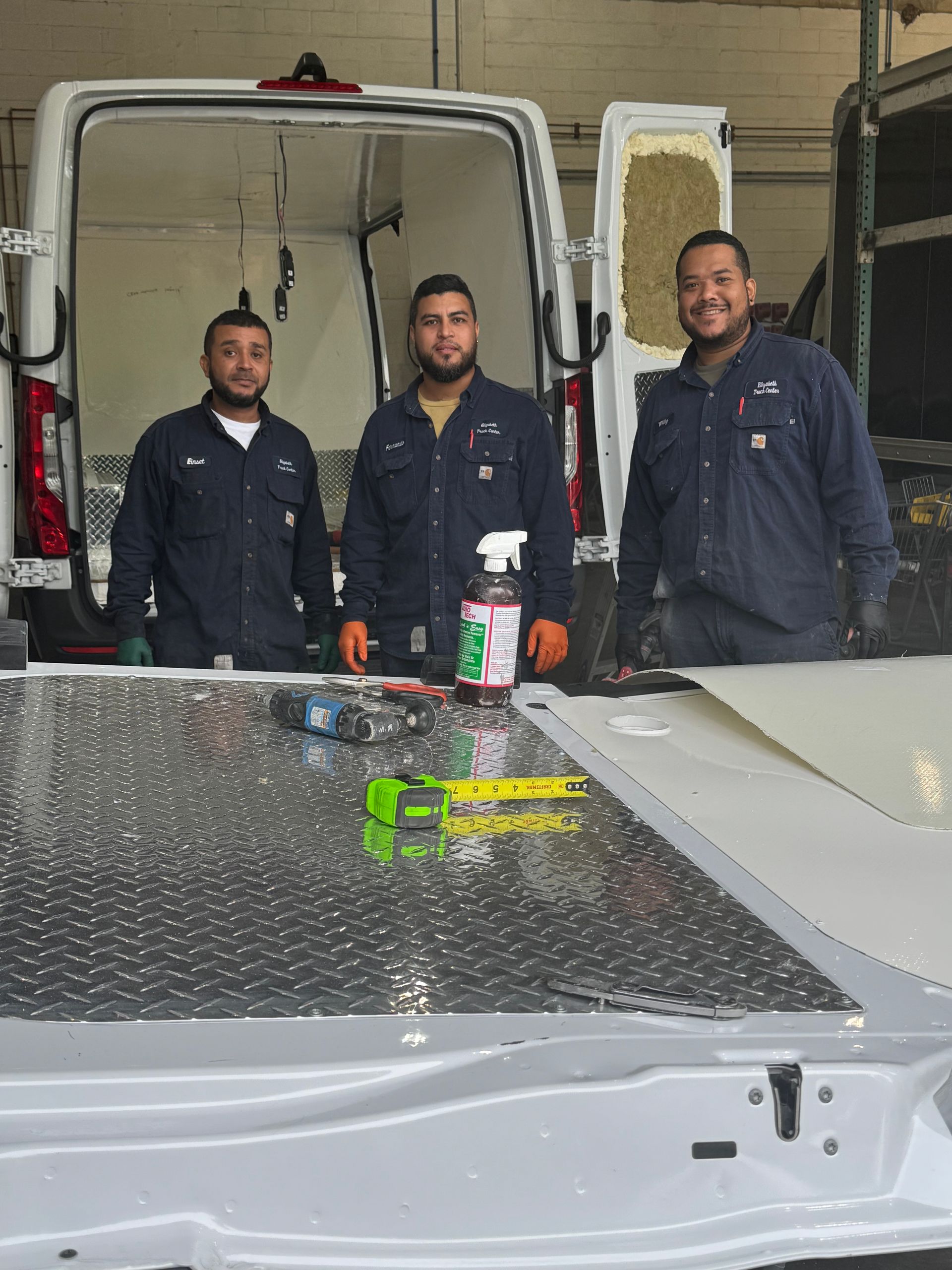 Three people wearing dark work uniforms stand in front of an open van, with tools and materials on a counter nearby.