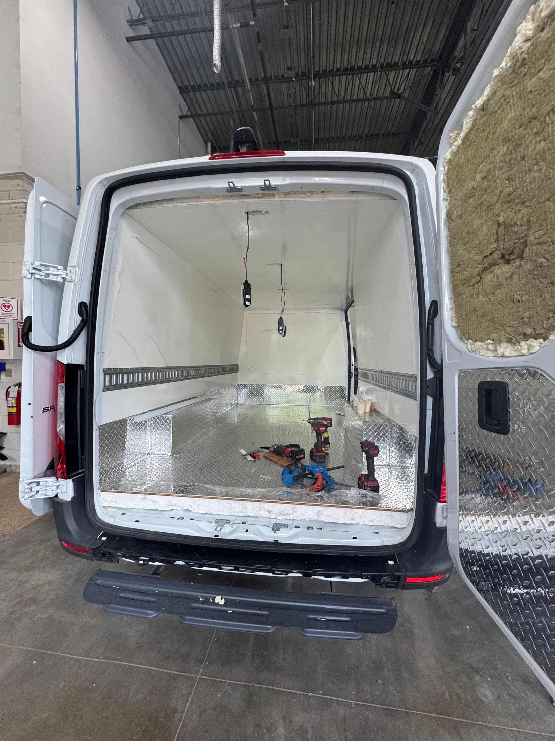 The open rear doors of a white cargo van reveal an empty, bright interior with metal flooring and partially insulated doors.