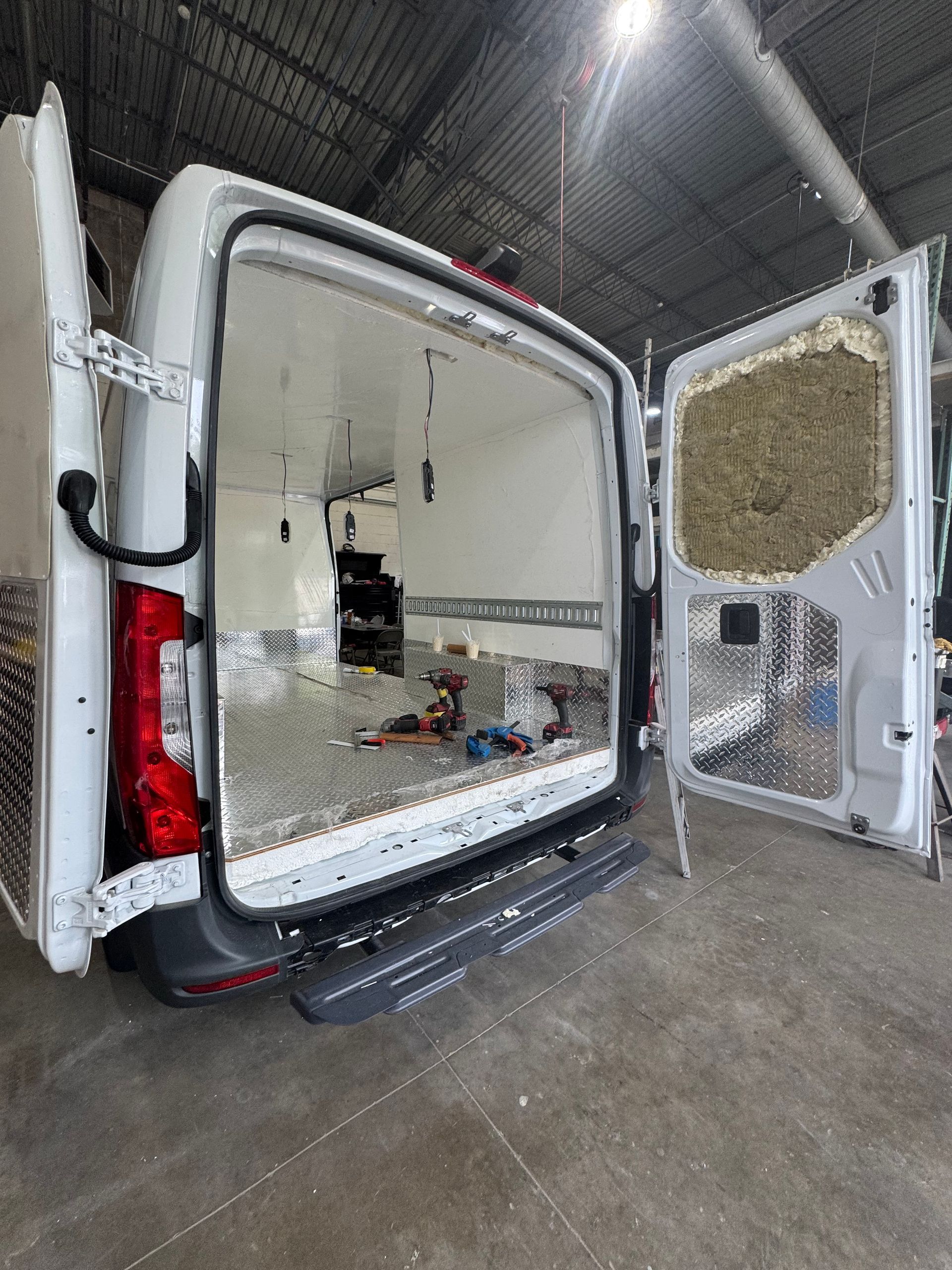 Rear view of an open white cargo van interior showing metal flooring, white-paneled walls, and insulation on the door.