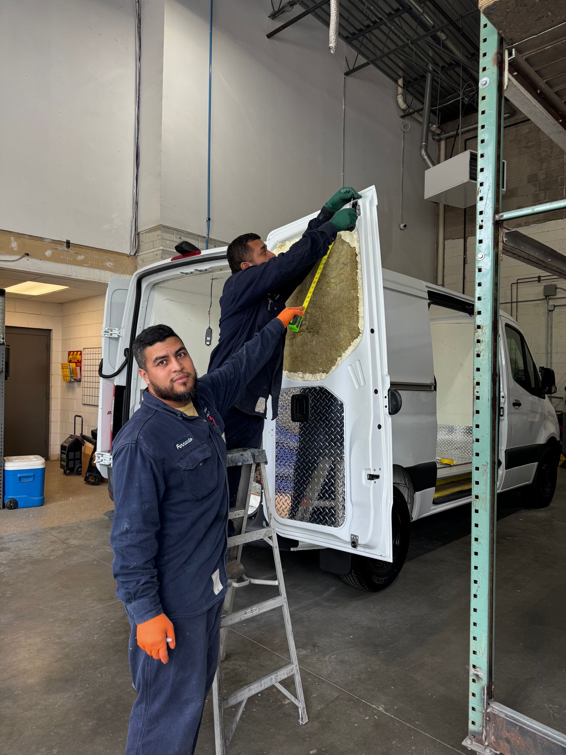 Two workers in blue uniforms install yellow insulation inside the rear door of a white cargo van in a workshop.