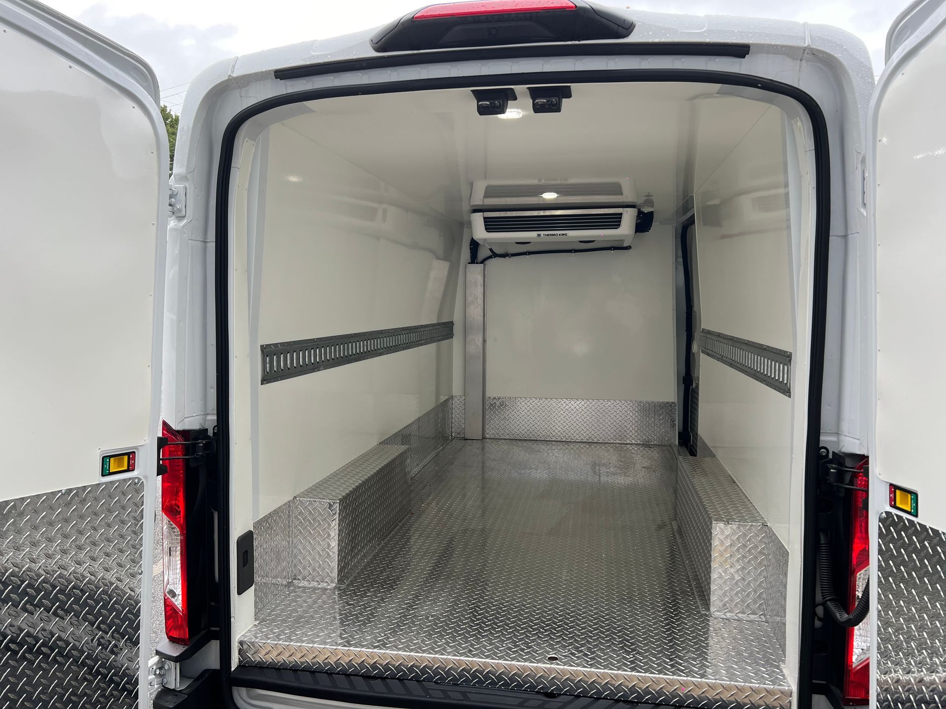 Rear view of a white refrigerated van with an open back, showing a metal-plated floor and cooling unit on the ceiling.