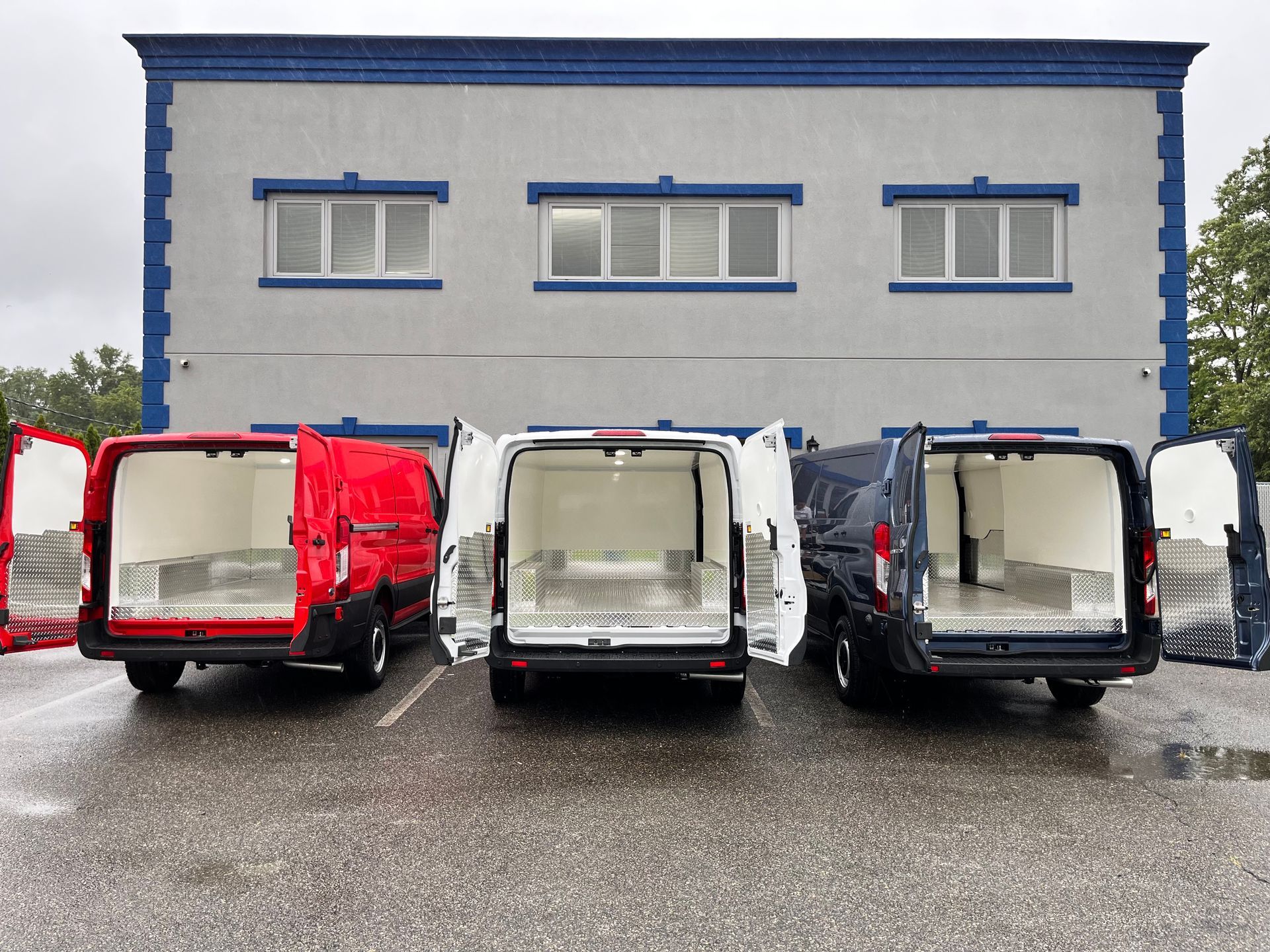 Three cargo vans in red, white, and dark blue are parked with their rear doors open in front of a light-colored building.