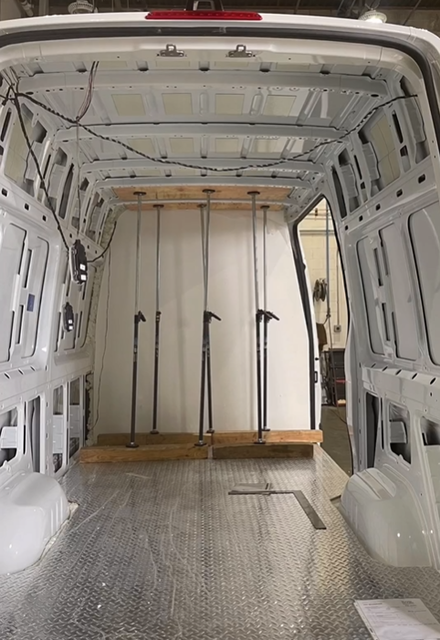 Interior of a cargo van featuring a diamond-plate metal floor, bare white walls, and four vertical cargo support poles.