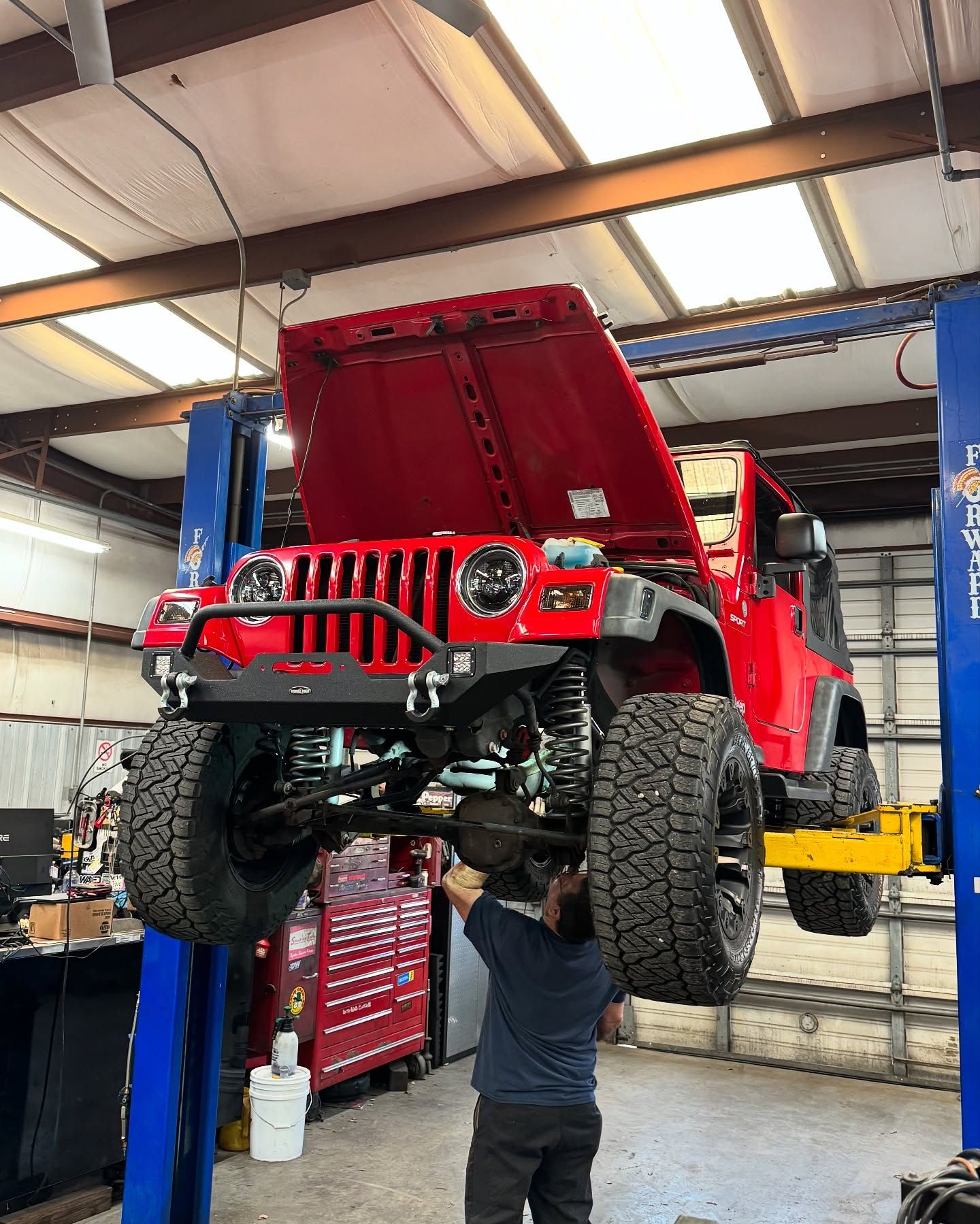 Red Jeep lifted in auto shop, mechanic working underneath with hood open