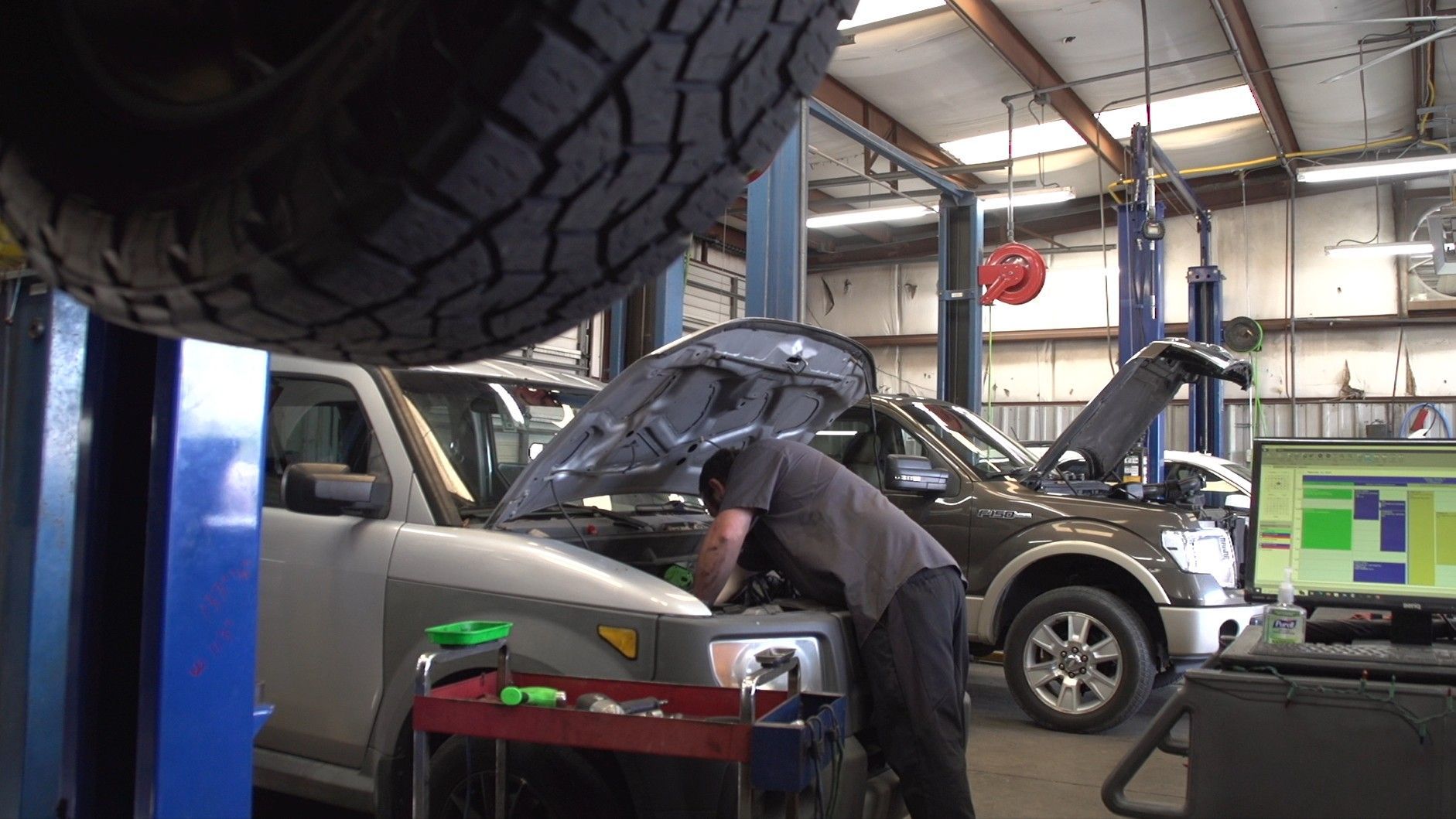 Auto repair shop with mechanic working on a silver car under a raised black truck tire