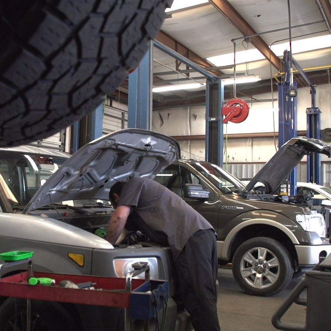 Mechanic working under the hood of a silver SUV in an auto repair shop with another car nearby